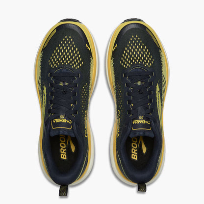 A pair of Brooks Cascadia 20 running shoes shown from a top-down view, featuring a dark navy blue and yellow mesh upper with a thick white midsole.