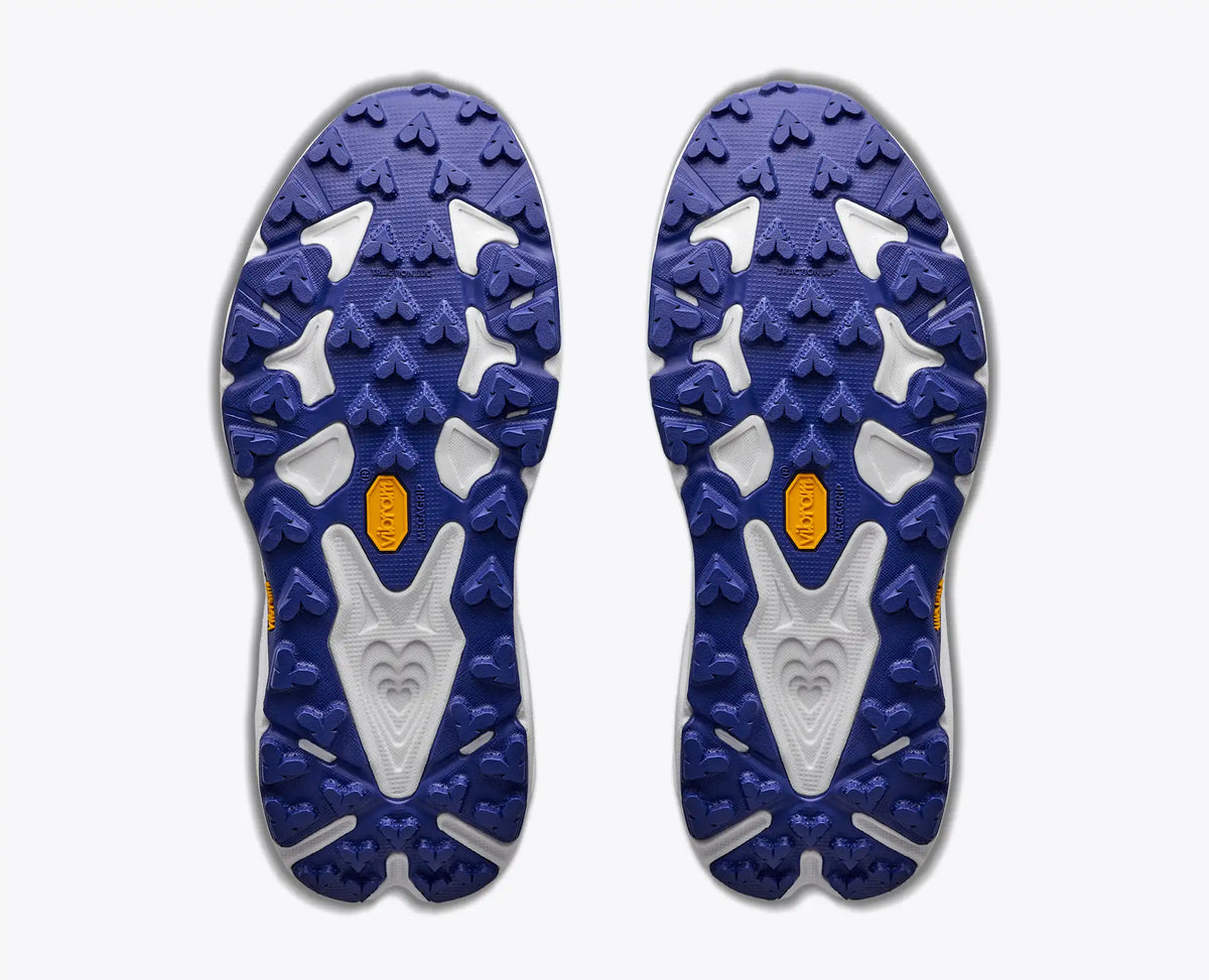 Top-down view of a pair of Hoka Speedgoat athletic shoe soles, showing a deep blue and white tread pattern with small orange Vibram logos, on a light gray background.