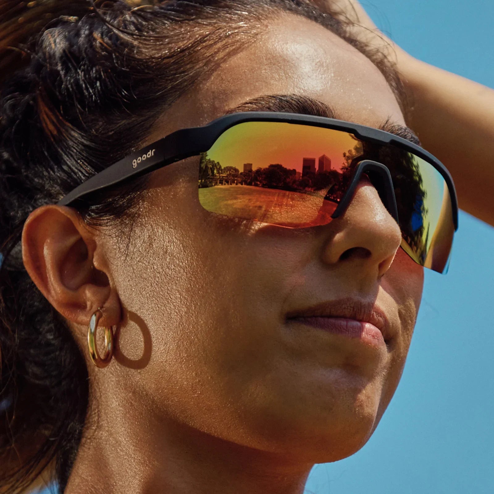 Person wearing reflective Goodr Bolt G: Do It For The Victory Dance sunglasses with a scenic reflection, against a clear blue sky.