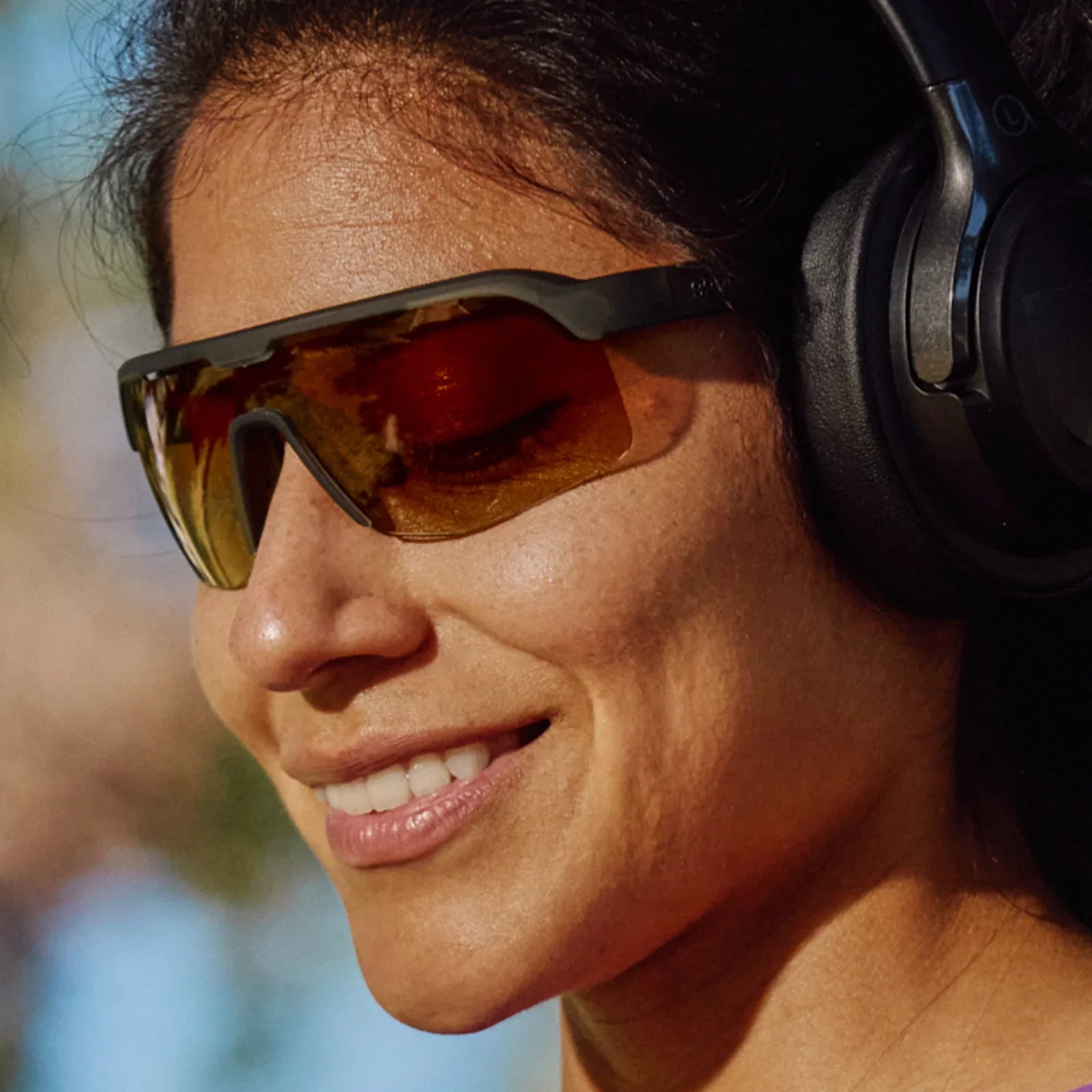 Woman wearing reflective Goodr Bolt G The Jungle Is My Gym wraparound sunglasses and large headphones with a blurred natural background