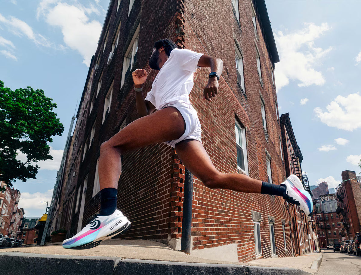 A man in a white t-shirt and white shorts running outdoors in white running shoes with pink, blue, and black accents, captured from a low angle against a brick building and blue sky.