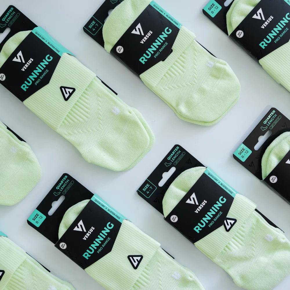 Multiple pairs of light yellow-green running socks in retail packaging, arranged diagonally on a white background. The packaging is black with teal and white text reading 'VERSUS RUNNING'.